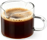 Clear glass creamer pitcher with two spouts and handle filled with coffee, white background