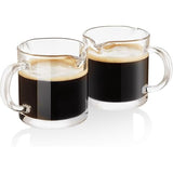 Two small glass espresso pitcher cups with dark coffee and crema on a white background